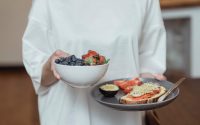 Person holding a bowl of fresh berries and a plate with avocado toast, displaying a healthy breakfast spread.